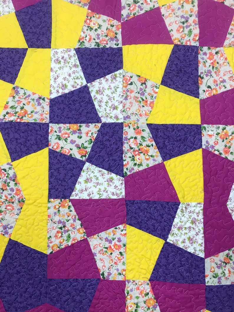 K&ouml;nnte beinhalten: Eine Steppdecke aus lila, gelbem und wei&szlig;em Stoff mit einem Blumenmuster. Die Steppdecke besteht aus vielen verschiedenen Formen und Gr&ouml;&szlig;en von Stoff.