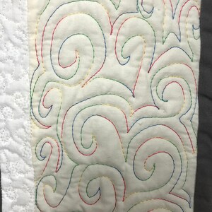 May include: A close-up of a white fabric with a decorative pattern of swirls stitched in red, blue, green, and gold thread.