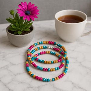 May include: Three beaded bracelets with turquoise, fuchsia, and gold beads. A white mug of tea and a potted plant with a pink flower are also in the image. The bracelets are on a marble surface.