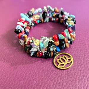 May include: Multi-colored beaded bracelet with a gold lotus charm. The bracelet is made of various stones, including black, red, blue, green, and brown.