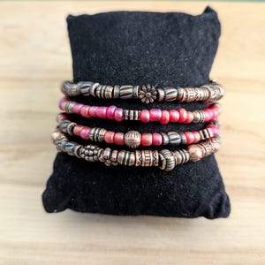 May include: A set of five bracelets with copper and red beads. The bracelets are stacked on a black velvet cushion.