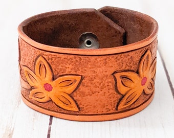 May include: A brown leather bracelet with a snap closure and a floral design. The flowers are orange with red centers.