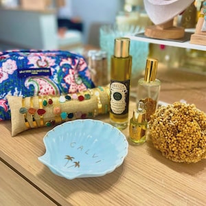 May include: A light blue ceramic shell-shaped dish with the text CALVI and two palm tree icons in gold, positioned near a floral print pouch, a burlap bracelet display, two glass bottles of Orelia oil, and a cluster of dried yellow flowers.