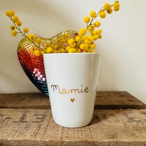 Può includere: Tazza in ceramica bianca con la parola "Mamie" scritta in caratteri dorati con un piccolo cuore dorato sotto. La tazza è piena di fiori di mimosa gialli.