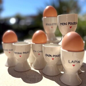 Peut inclure: Plusieurs coquetiers en céramique blanche, chacun contenant un œuf brun. Les tasses sont inscrites avec du texte noir et un cœur rouge. Le texte comprend "Maman Poule", "Papa Poule" et "Lapin".