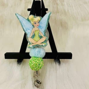 May include: A badge reel featuring a cartoon fairy with light blue glitter wings and a green dress. The reel has a light blue pom-pom and a green glitter ball. The badge reel is attached to a silver clip.