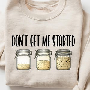 May include: Beige sweatshirt with the phrase "DON'T GET ME STARTED" in black. Below the text are three jars with yellow contents. The sweatshirt has long sleeves and a crew neck.