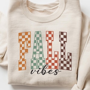 May include: A cream-coloured sweatshirt featuring the words "FALL vibes" in a checkered design. The letters are in orange, green, red, and brown. The sleeves are rolled up. The sweatshirt is laid flat.