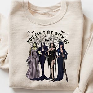 May include: Beige sweatshirt featuring a graphic of four women in gothic attire, with the text "YOU CAN'T SIT WITH US" above them. The design includes bats and a spiderweb. The sweatshirt is folded on a white surface.