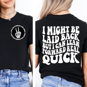 May include: Black t-shirt with white text on the front and back. The front features a skeleton hand making a peace sign with the text "I CAN LEAN FORWARD REAL QUICK" around it. The back says "I MIGHT BE LAID BACK BUT I CAN LEAN FORWARD REAL QUICK."