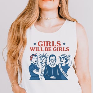 May include: White tank top with the text "GIRLS WILL BE GIRLS" in red and blue. The graphic features illustrations of four women in blue, including Rosie the Riveter, the Statue of Liberty, Ruth Bader Ginsburg, and a woman with a "VOTE FOR WOMEN" sash.