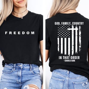 May include: Black t-shirt with the word "FREEDOM" in white block letters on the front. The back of the shirt features an American flag design with a cross and the text "GOD, FAMILY, COUNTRY IN THAT ORDER - CHARLIE KIRK."