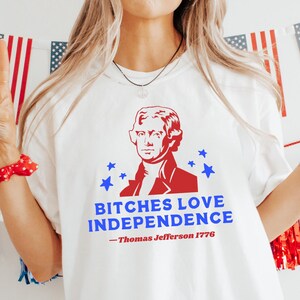 May include: White t-shirt with a red and blue graphic of Thomas Jefferson and the text "BITCHES LOVE INDEPENDENCE - Thomas Jefferson 1776". The image is decorated with American flags and red, white, and blue decorations.