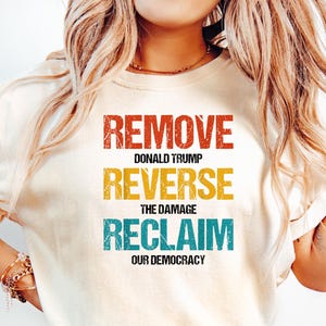 May include: A white t-shirt with a vintage-style graphic that reads "Remove Donald Trump, Reverse the Damage, Reclaim Our Democracy".