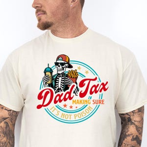 Pode incluir: Camiseta creme com um gr&aacute;fico circular. O gr&aacute;fico apresenta o texto "Dad Tax" em vermelho, um esqueleto usando chap&eacute;u e &oacute;culos de sol, segurando uma bebida e batatas fritas, e a frase "Making Sure It's Not Poison".