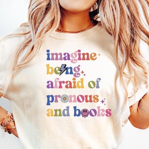 May include: A white t-shirt with the text "imagine being afraid of pronouns and books" in colorful letters with a lightning bolt, daisy, and smiley face graphic.