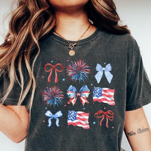 May include: A dark gray t-shirt with a graphic print of red, white, and blue bows, fireworks, and American flags.