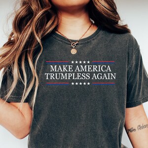 May include: A dark gray t-shirt with the text "MAKE AMERICA TRUMPLESS AGAIN" printed on the front. The text is in white with red and blue stars above and below it.