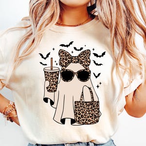 May include: Cream-colored t-shirt featuring a Halloween ghost design. The ghost wears sunglasses, a leopard print bow, and carries a leopard print tote bag and a coffee cup. Black bats are scattered around the ghost.