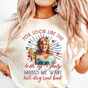 May include: A white t-shirt with a red, white, and blue graphic featuring a woman holding a hot dog. The graphic says "You look like the 4th of July makes me want hot dog real bad."