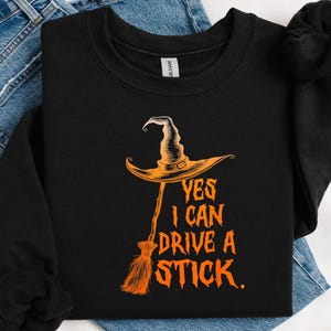 May include: Black crewneck sweatshirt with an orange witch hat and broom graphic. The text "YES I CAN DRIVE A STICK" is printed in orange. The sweatshirt is folded on top of blue jeans.