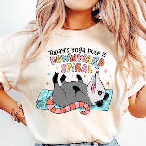 May include: Cream-colored t-shirt with the text "Today's yoga pose is DOWNWARD SPIRAL" and a cartoon opossum on a yoga mat. The opossum is gray with a pink and orange tail. The shirt has a relaxed fit.