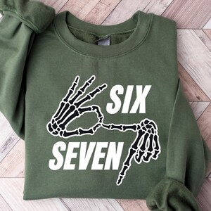 May include: An olive green sweatshirt with the words "SIX SEVEN" in white, accompanied by a black and white skeleton hand graphic. The sweatshirt is folded on a light wood-grain surface.