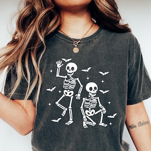 May include: Dark gray t-shirt featuring a white graphic of two skeletons and bats, perfect for Halloween. The skeletons are in playful poses, with one waving and the other squatting. The shirt is made of a soft material.