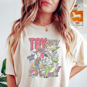 Puede incluir: Camiseta color crema con un gr&aacute;fico colorido de los personajes de Toy Story, incluyendo a Woody, Buzz Lightyear y Rex. El dise&ntilde;o incluye el texto "Toy Story" y "Since 1995". Una prenda de vestir casual.