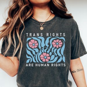 May include: A dark grey t-shirt with the text "TRANS RIGHTS ARE HUMAN RIGHTS" printed in white. The design features three pink flowers with blue centres and blue leaves on a wavy blue background.