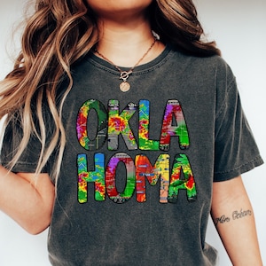 May include: A dark gray t-shirt with a colorful graphic design that reads "OKLA HOMA". The graphic is made up of a weather radar map.