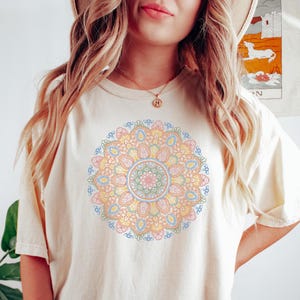 May include: A cream colored t-shirt with a colorful mandala design printed on the front. The mandala is in shades of pink, yellow, blue, and green.