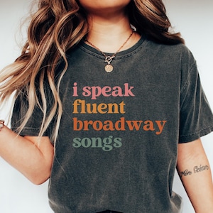 May include: Dark gray t-shirt with the text "i speak fluent Broadway songs" in pink, orange, and green. The shirt is paired with blue jeans and white sneakers. A gold necklace and bracelet are also visible.
