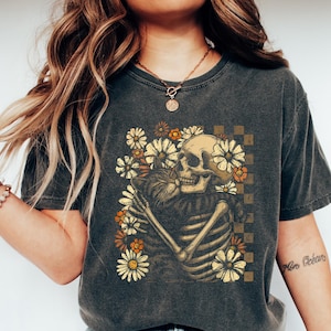 May include: Dark gray t-shirt featuring a skeleton embracing a cat, surrounded by flowers. The design includes a checkered background. The shirt is worn with blue jeans and white slip-on shoes. The overall theme is a blend of gothic and floral elements.