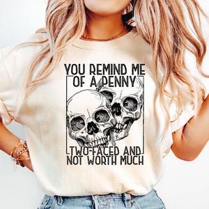 May include: Cream-colored t-shirt featuring a black and white graphic of three skulls within a rectangular frame. The text above reads "YOU REMIND ME OF A PENNY," and below, "TWO-FACED AND NOT WORTH MUCH."