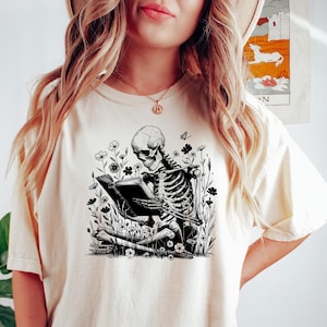 May include: Cream-colored t-shirt featuring a black and white illustration of a skeleton reading a book amidst flowers. The design is centered on the shirt, creating a unique graphic tee.