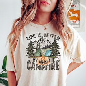 May include: A white t-shirt with a graphic print of a mountain scene with a campfire, tents, and trees. The text "Life is better by the campfire" is printed on the shirt.