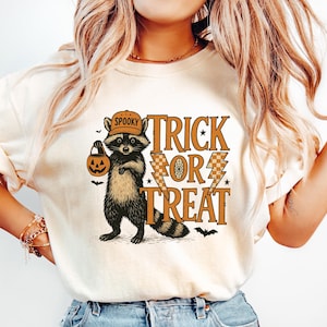 Peut inclure: T-shirt cr&egrave;me avec un motif d'Halloween. Le design comprend un raton laveur portant un chapeau et tenant une lanterne citrouille, avec les mots "Spooky Trick or Treat" en orange et marron.