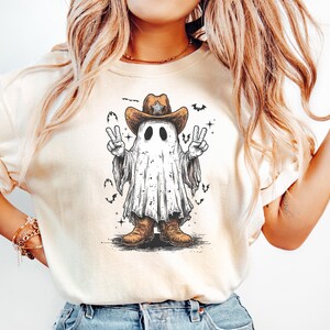 May include: Cream-colored t-shirt featuring a cartoon ghost wearing a cowboy hat and boots. The ghost is making a peace sign with both hands. Bats and stars are also in the design.