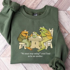 Puede incluir: Sudadera verde con una ilustraci&oacute;n de dibujos animados de dos ranas en una mesa. Las ranas visten ropa y est&aacute;n comiendo. El texto de la sudadera dice: "&iexcl;Debemos dejar de comer!" grit&oacute; Sapo mientras com&iacute;a otro.