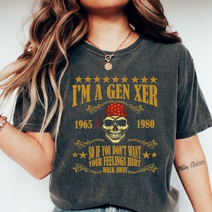 Op de afbeelding: Een donkergrijs T-shirt met een vintage-stijl grafische afbeelding met een schedel die een rode bandana draagt en de tekst "I'm a Gen Xer 1965 - 1980 So if you don't want your feelings hurt walk away".