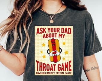Puede incluir: Una camiseta gris oscuro con un gráfico de un hot dog con cara y brazos. El texto en la camiseta dice "Ask your dad about my throat game. Downing Daddy's special sauce."