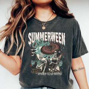 Puede incluir: Camiseta gris oscuro con las palabras "SUMMERWEEN Vibes" en una fuente desgastada. El gr&aacute;fico presenta un esqueleto relaj&aacute;ndose en una silla de playa bajo una sombrilla, con un gato negro y murci&eacute;lagos. Tambi&eacute;n se incluye el texto "Spooky Year Round".