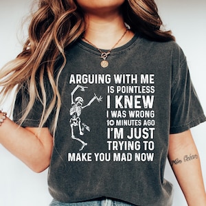 May include: Dark gray t-shirt with the text "ARGUING WITH ME IS POINTLESS I KNEW I WAS WRONG 10 MINUTES AGO I'M JUST TRYING TO MAKE YOU MAD NOW" and a skeleton graphic. The shirt is paired with blue jeans and white shoes.