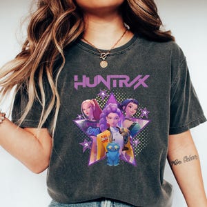 May include: Dark gray t-shirt featuring a graphic of three animated women and a blue cat, with the word "HUNTRAX" in purple. The design is set against a star-shaped background.