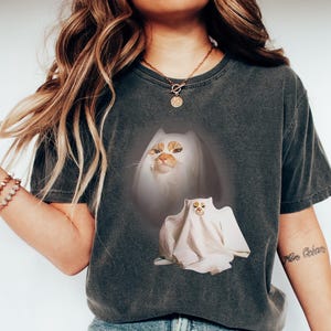 Puede incluir: Camiseta gris oscuro con un dise&ntilde;o de gato. El gr&aacute;fico muestra un gato con pelaje naranja y blanco, y un segundo gato disfrazado de fantasma. La camiseta es de estilo c&oacute;modo e informal.