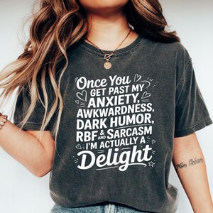 Pu&ograve; includere: Una maglietta grigia scura con testo bianco che dice "Once You Get Past My Anxiety, Awkwardness, Dark Humor, RBF & Sarcasm I'm Actually A Delight".
