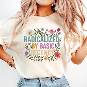 May include: Cream-colored t-shirt with the text "RADICALIZED BY BASIC DECENCY" in green, purple, and yellow, surrounded by a floral design. The model is wearing light blue jeans.