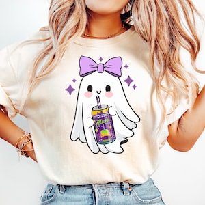 K&ouml;nnte beinhalten: Cremefarbenes T-Shirt mit einem niedlichen Geister-Cartoon, der eine lila Schleife tr&auml;gt und eine Dose "Witch's Brew"-Limonade h&auml;lt. Der Geist l&auml;chelt, und es gibt lila Sternakzente. Das Shirt wird mit hellblauen Jeans getragen.