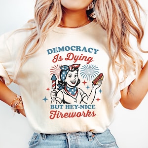 May include: Cream-colored t-shirt with a graphic featuring a woman holding a firecracker and a hot dog, with text that reads "DEMOCRACY Is Dying BUT HEY-NICE FIREWORKS" in red and blue. The design has a vintage, patriotic aesthetic.